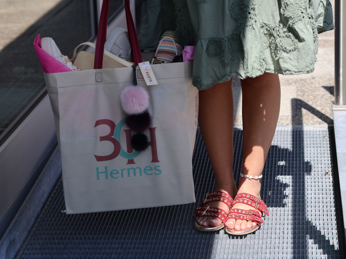 Frau im grünen Sommerkleid mit roten Sandalen und Hermes-Einkaufstasche mit Pompon-Anhänger voller Accessoires am Eingang