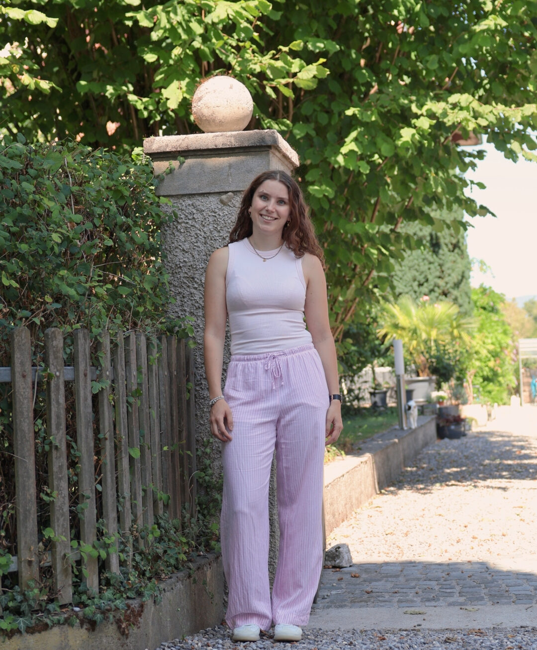 Frau in rosa Hose und hellrosa Top lächelt auf Kiesweg im grünen Garten – sommerlicher Casual Look