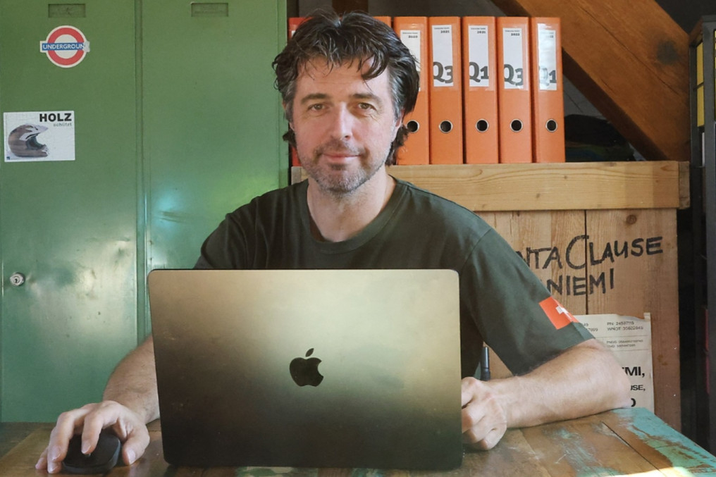 Mann arbeitet am MacBook im Homeoffice-Büro, sitzt am Holztisch mit Maus; Ordnerregal und Werkstattflair im Hintergrund