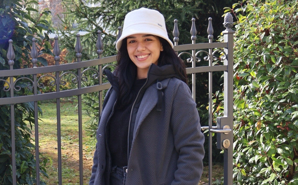 Lächelnde Frau mit Bucket Hat und grauem Mantel vor schmiedeeisernem Gartentor im Grünen, herbstliches Outdoor-Porträt.